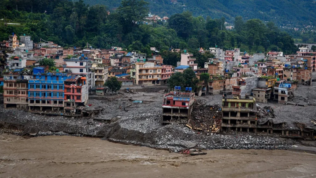 Flood-damaged riverside town in Nepal with muddy, debris-filled water in the foreground and densely packed colorful buildings beneath green hills.