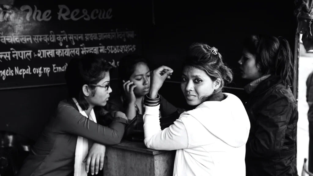 Black-and-white backstage scene: women gathered around a booth, one looks toward the camera, with Nepali writing on a chalkboard behind them.