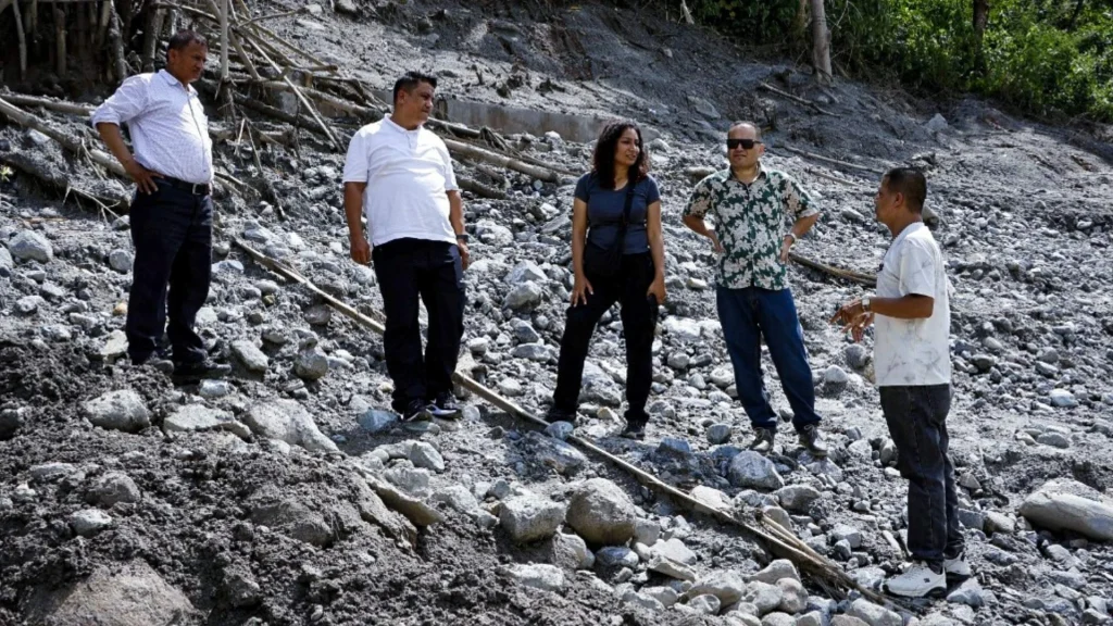 Five people stand on a steep, rocky landslide slope in Nepal, discussing the rugged terrain amid fallen branches and forest, conveying concern.