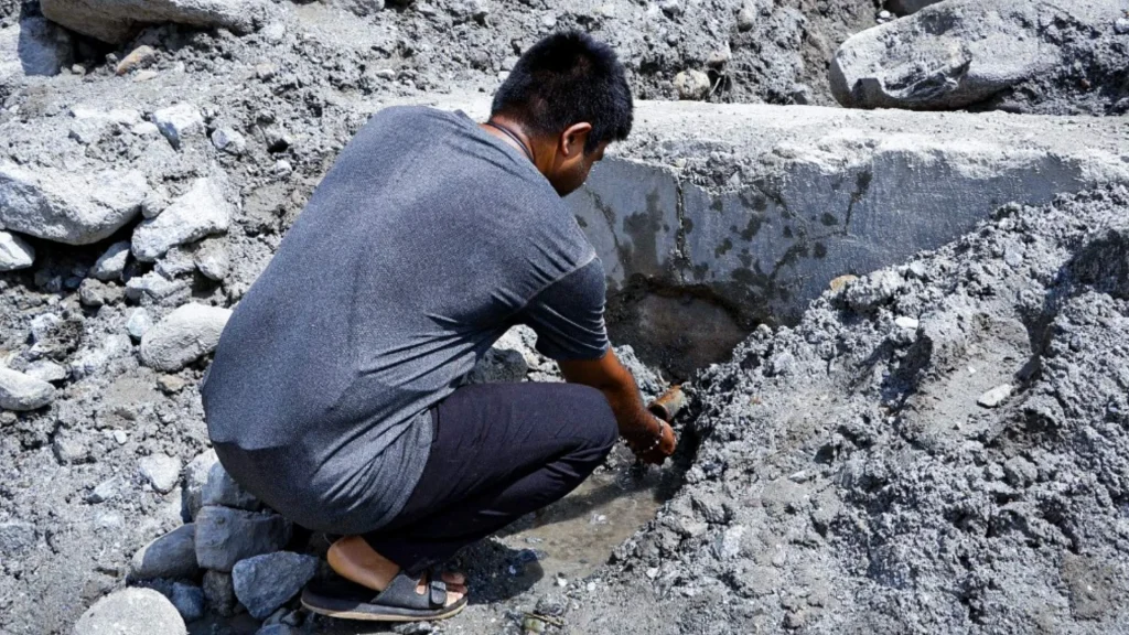 A man in dark clothing crouches among gray rocks in Nepal, examining debris beside a weathered concrete barrier in a stark landscape.