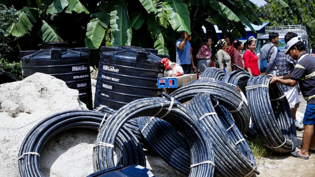 Large coils of black electrical cable lie beside sand and water tanks, while workers gather at a busy outdoor construction site under banana trees in Nepal.