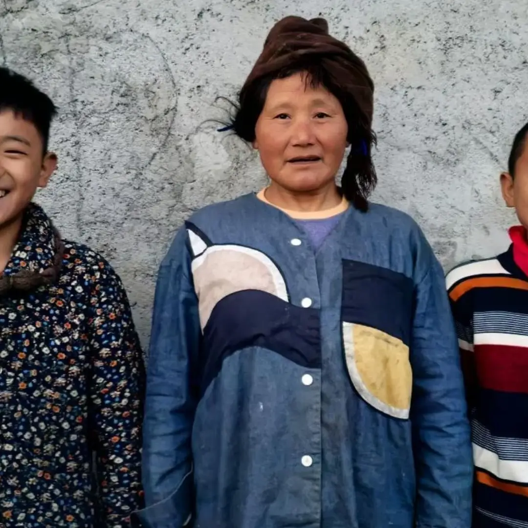 A smiling boy in a floral jacket, a woman in a blue shirt with a head wrap, and a boy in a striped sweater with a red scarf stand by a gray wall in rural China.