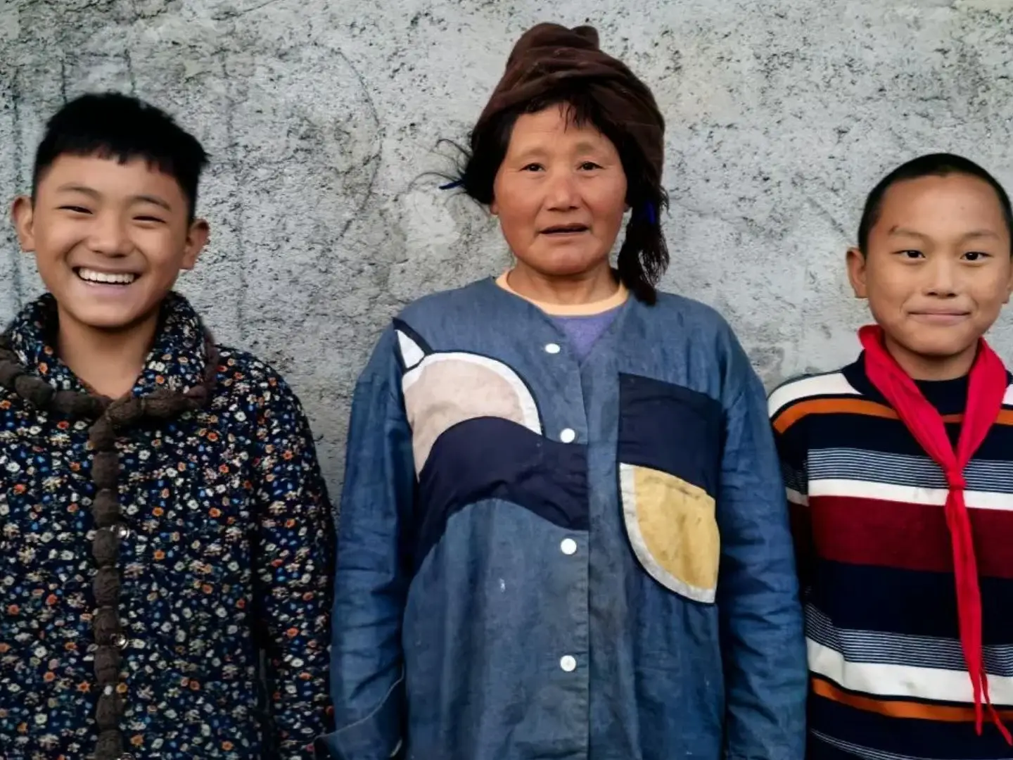 A smiling boy in a floral jacket, a woman in a blue shirt with a head wrap, and a boy in a striped sweater with a red scarf stand by a gray wall in rural China.