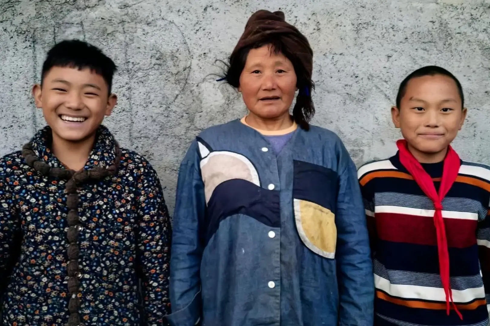 A smiling boy in a floral jacket, a woman in a blue shirt with a head wrap, and a boy in a striped sweater with a red scarf stand by a gray wall in rural China.