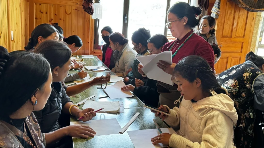 A group of women sit at a long table in a wood-paneled room, working with papers, pens, and a smartphone during a collaborative workshop session in rural China.
