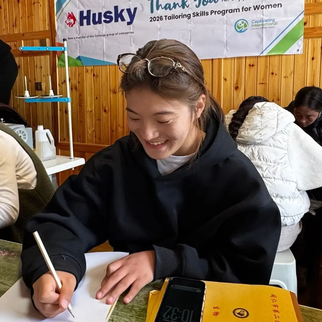 A young woman in a black hoodie writes on paper at a table in a room with wood-paneled walls, surrounded by others in a tailoring skills program in rural China.
