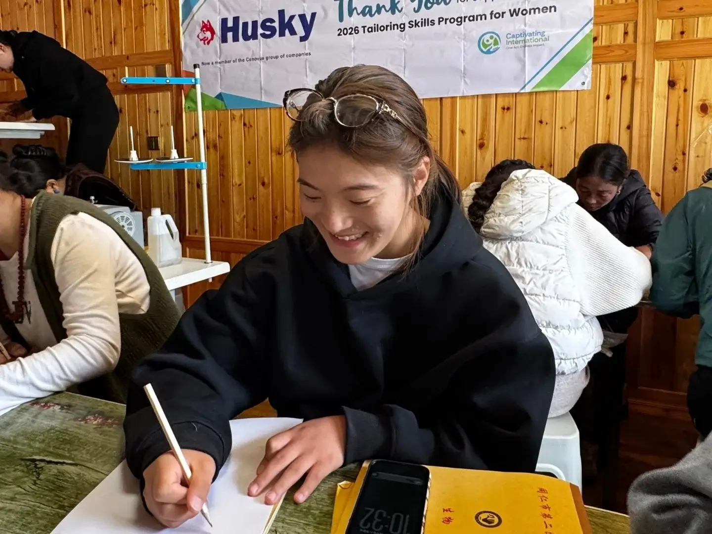 A young woman in a black hoodie writes on paper at a table in a room with wood-paneled walls, surrounded by others in a tailoring skills program in rural China.