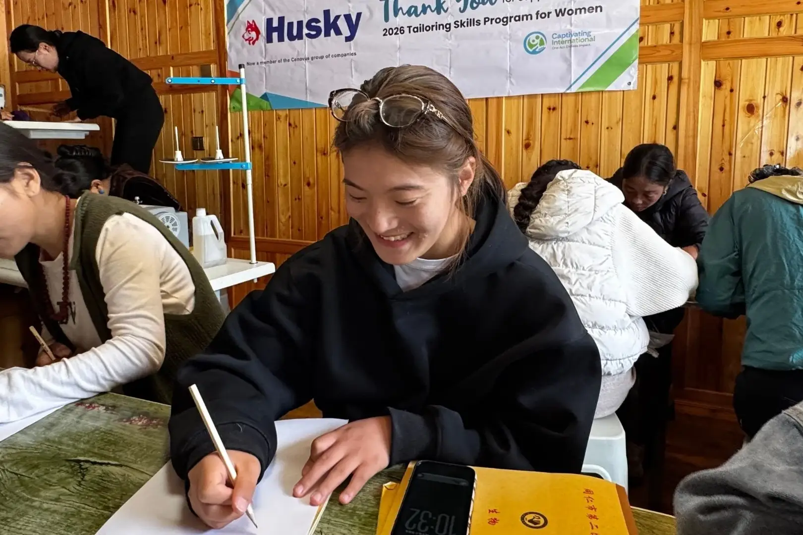 A young woman in a black hoodie writes on paper at a table in a room with wood-paneled walls, surrounded by others in a tailoring skills program in rural China.