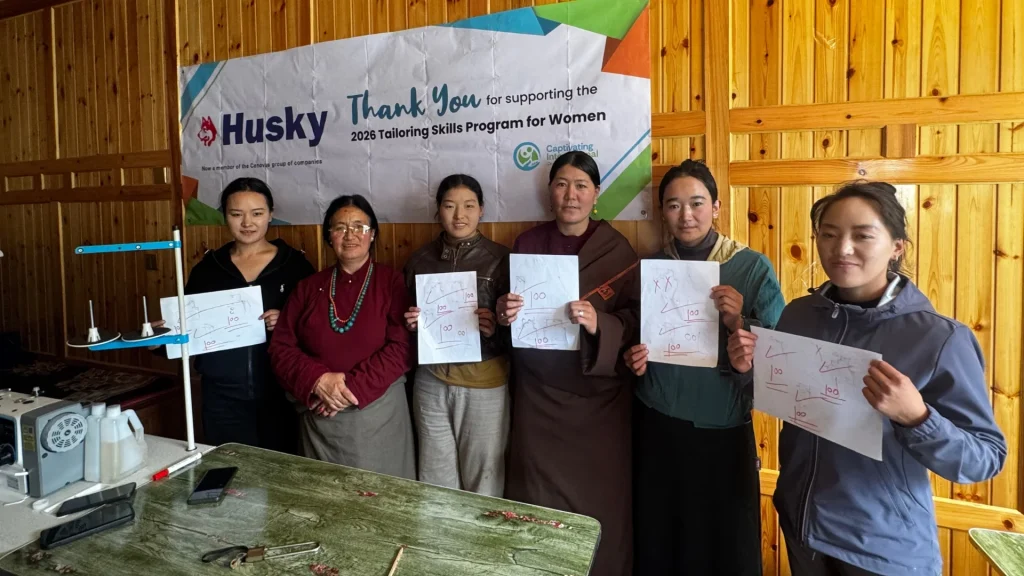 Six women stand in front of a banner, holding up papers with handwritten notes and scores, in a room with wood-paneled walls and a sewing table in rural China.
