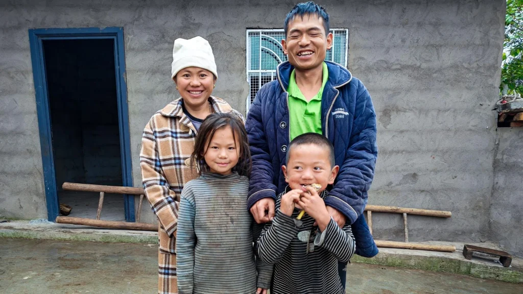 Smiling family of four poses outside a simple concrete home in rural China: parents stand behind two children in a rural setting, creating a warm, joyful mood.