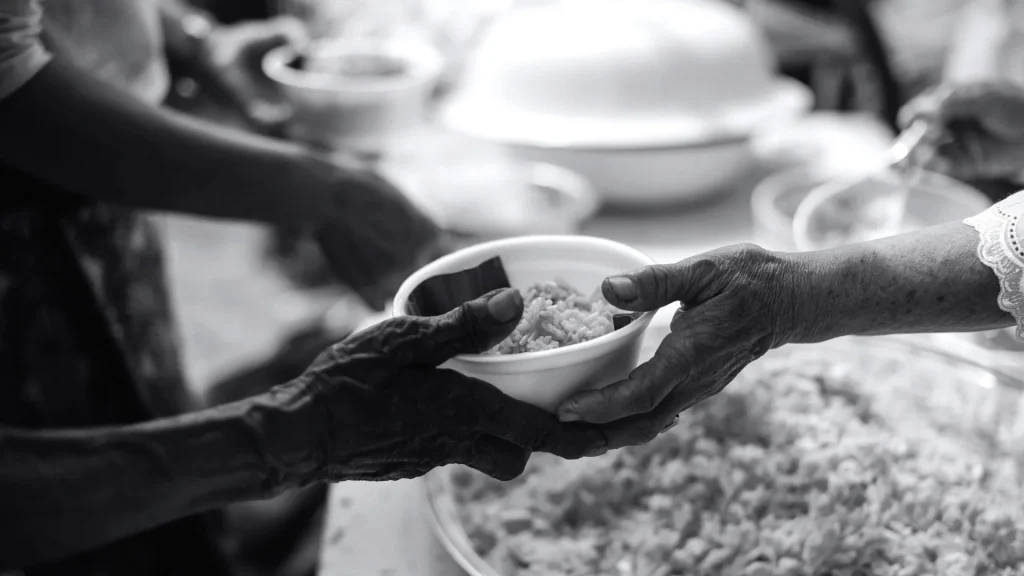 Black-and-white photo of hands passing a bowl of rice at a communal meal, conveying generosity, care, and shared nourishment.