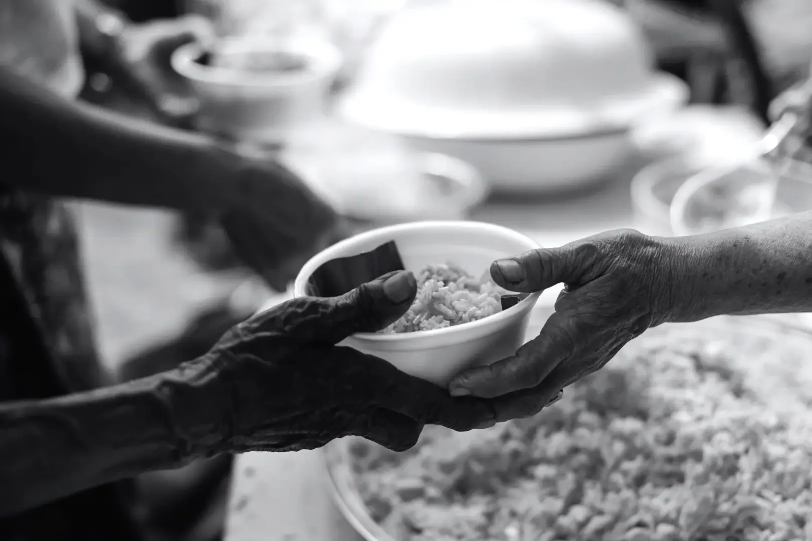 Black-and-white photo of hands passing a bowl of rice at a communal meal, conveying generosity, care, and shared nourishment.