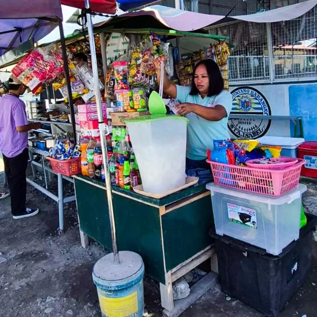 A woman stands behind a street drink stand, serving beverages to customers in a bustling urban environment in the Philippines.