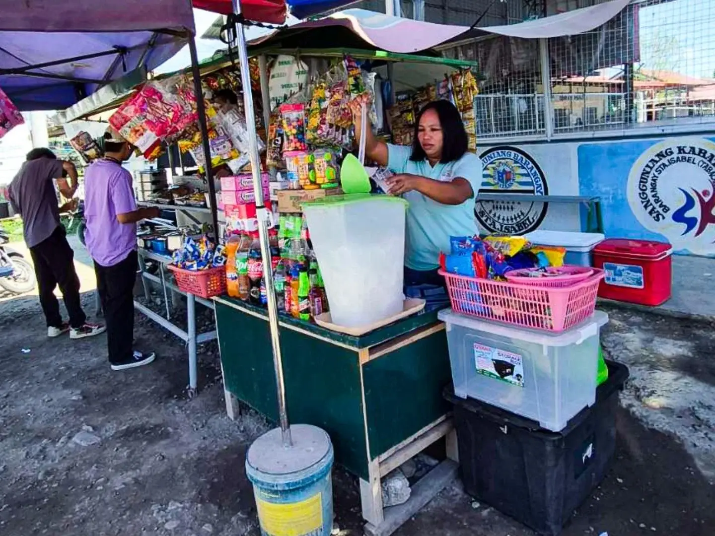 A woman stands behind a street drink stand, serving beverages to customers in a bustling urban environment in the Philippines.