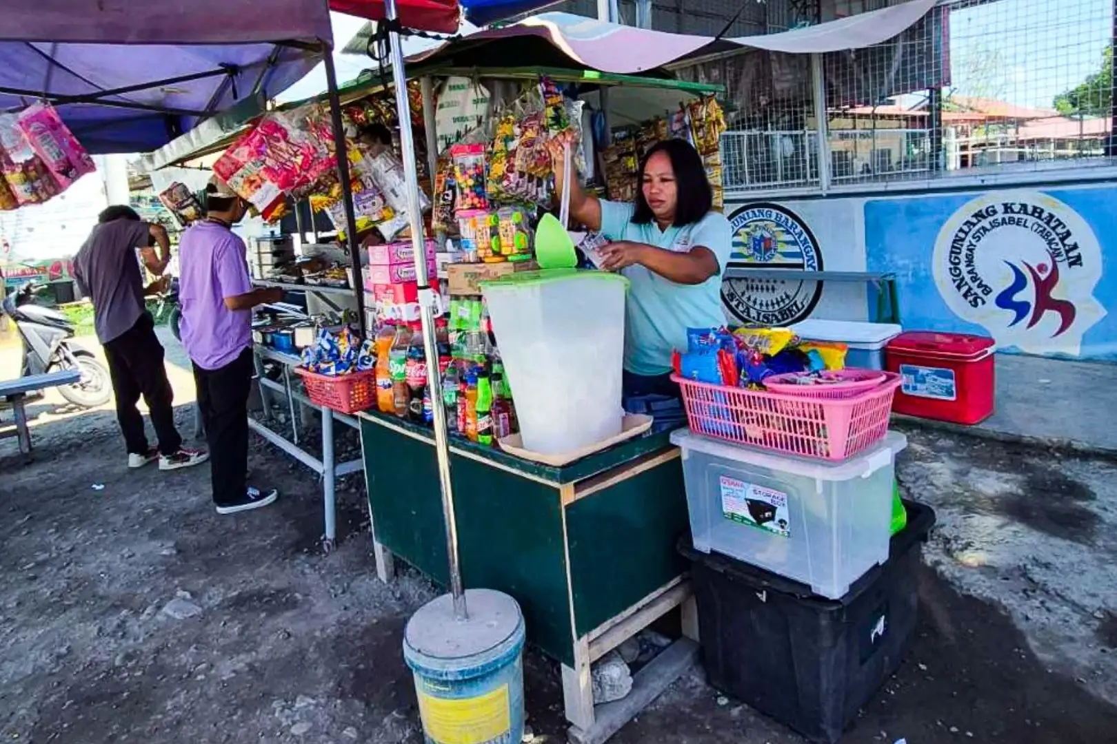 A woman stands behind a street drink stand, serving beverages to customers in a bustling urban environment in the Philippines.