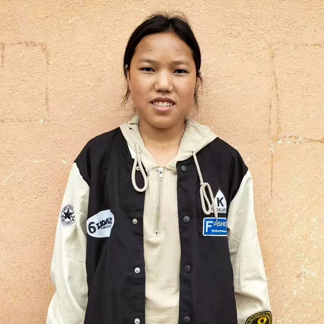A young woman in a jacket stands against a wall in rural China.