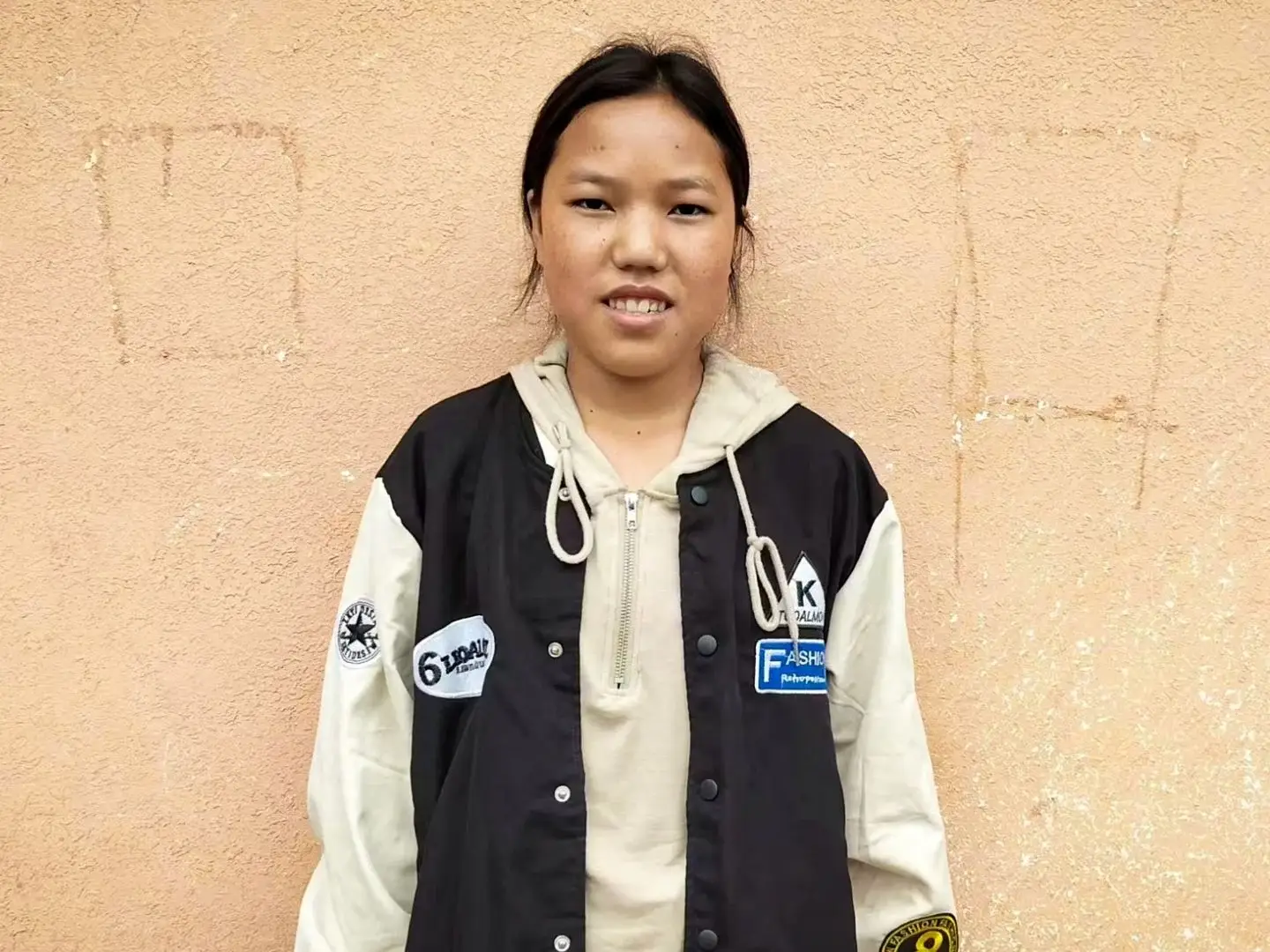 A young woman in a jacket stands against a wall in rural China.