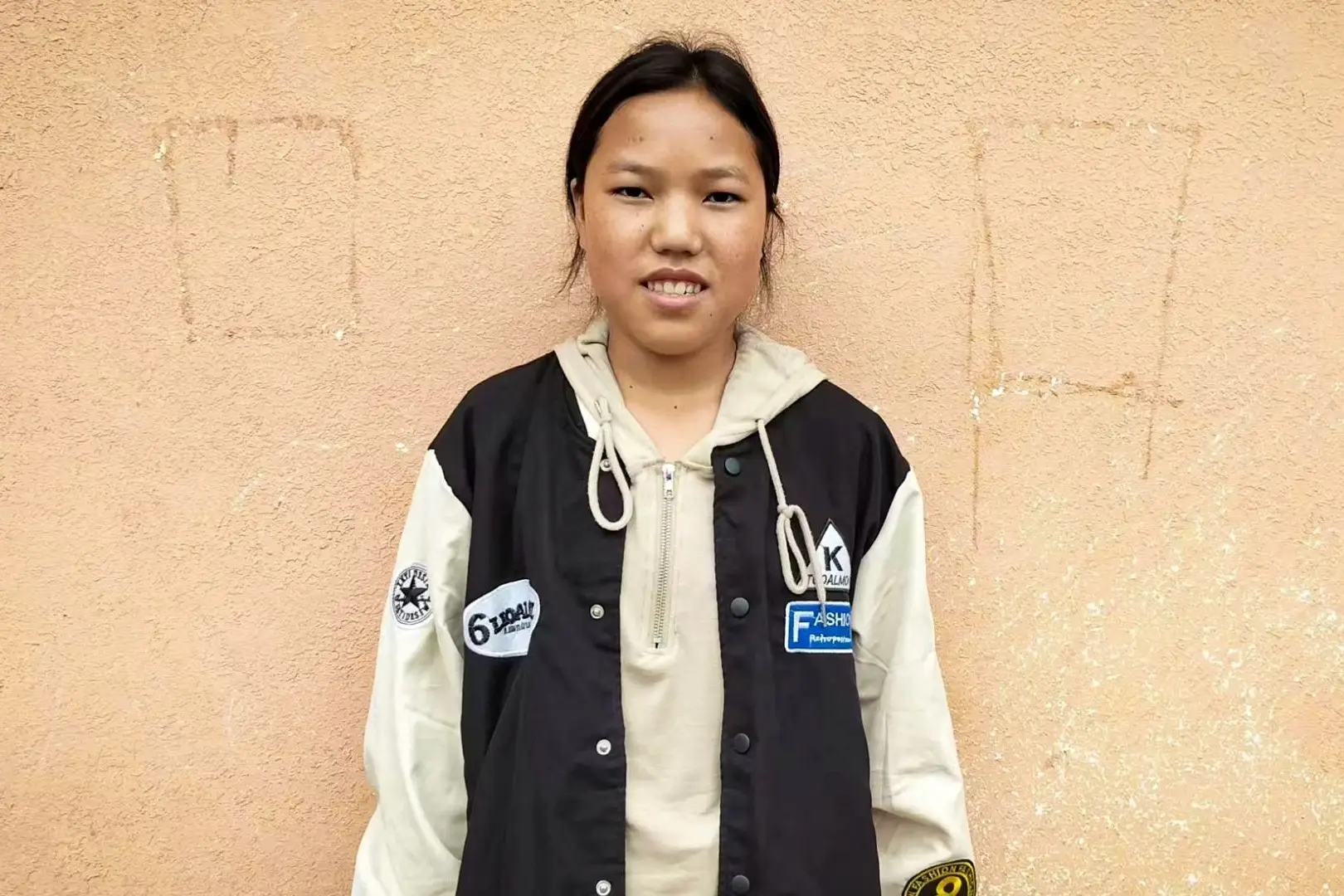 A young woman in a jacket stands against a wall in rural China.