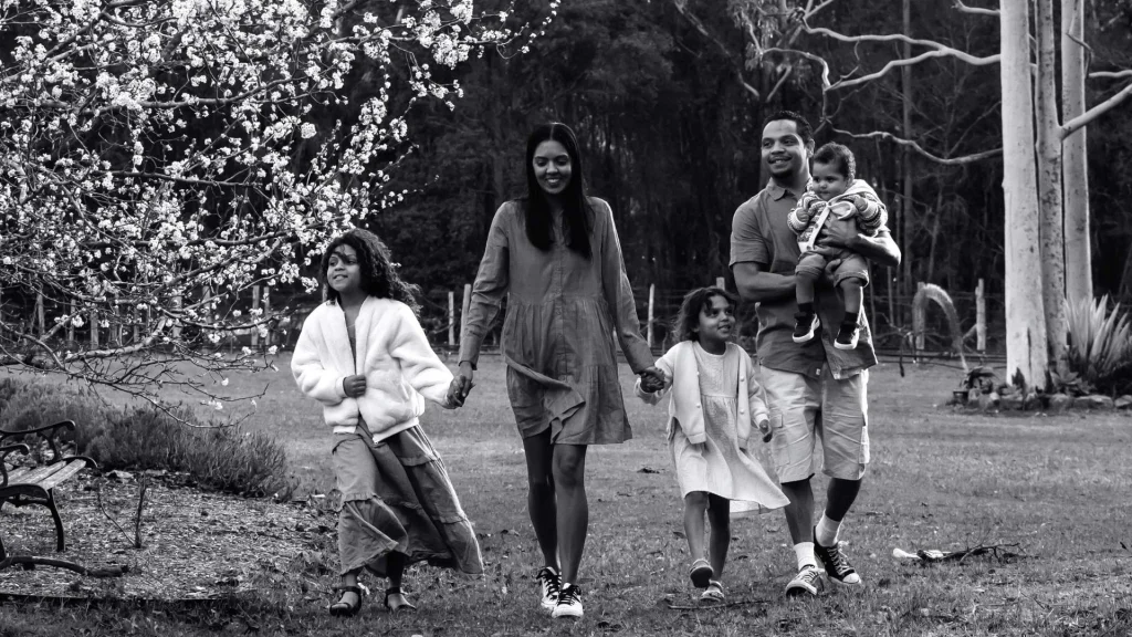 Black-and-white photo of a joyful family walking hand in hand through a wooded park, with blooming trees, grass, and tall trunks behind them.