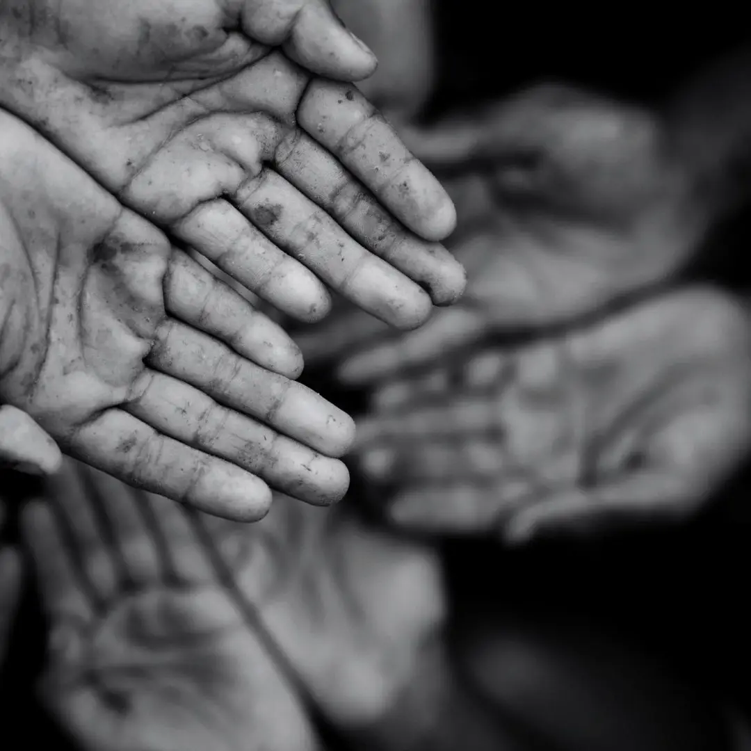 Black-and-white close-up of several outstretched, dirt-streaked hands reaching toward the camera, evoking hardship, vulnerability, and a plea for help.
