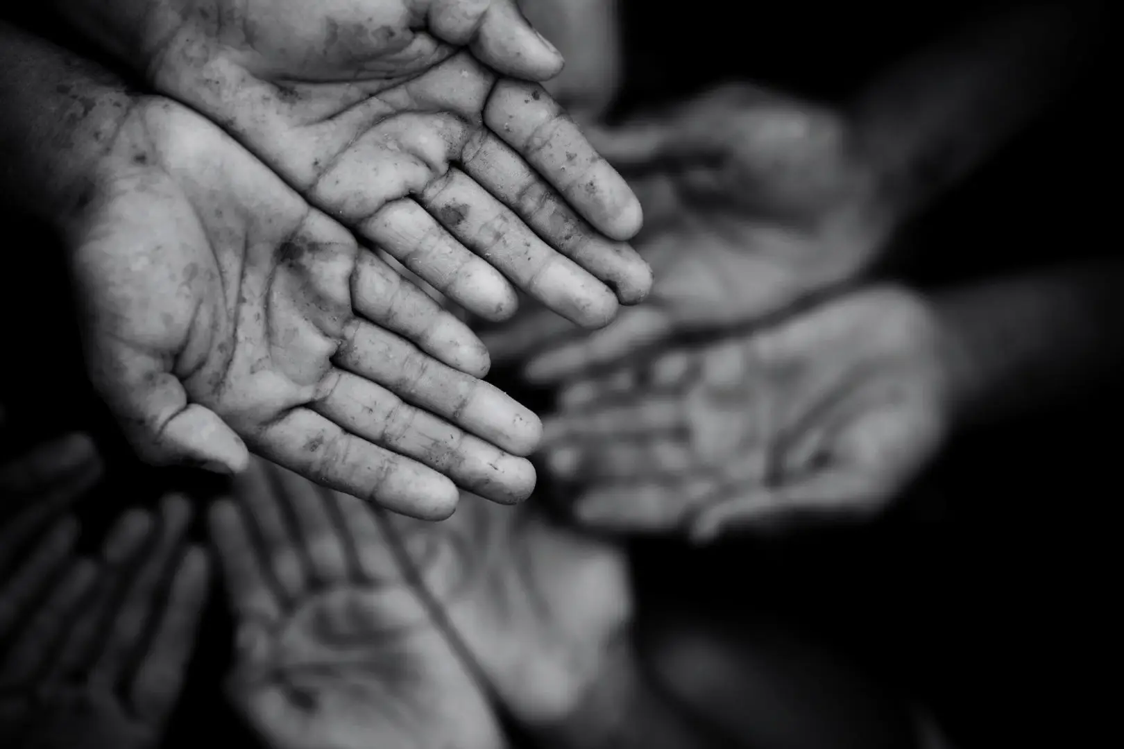 Black-and-white close-up of several outstretched, dirt-streaked hands reaching toward the camera, evoking hardship, vulnerability, and a plea for help.