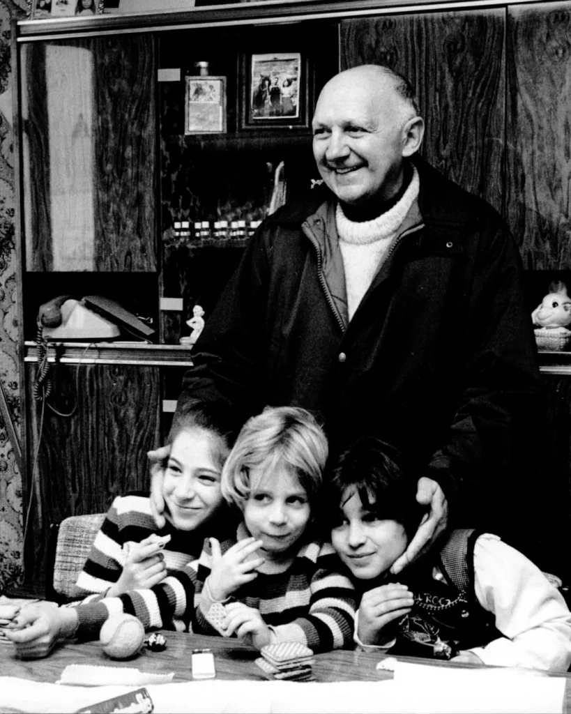 A smiling bald man stands behind three children gathered at a table in a wood-paneled room; the black-and-white portrait feels warm and affectionate.