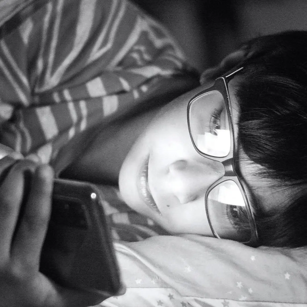 A boy wearing glasses lies in bed, holding a cell phone and looking at the screen with a focused expression.
