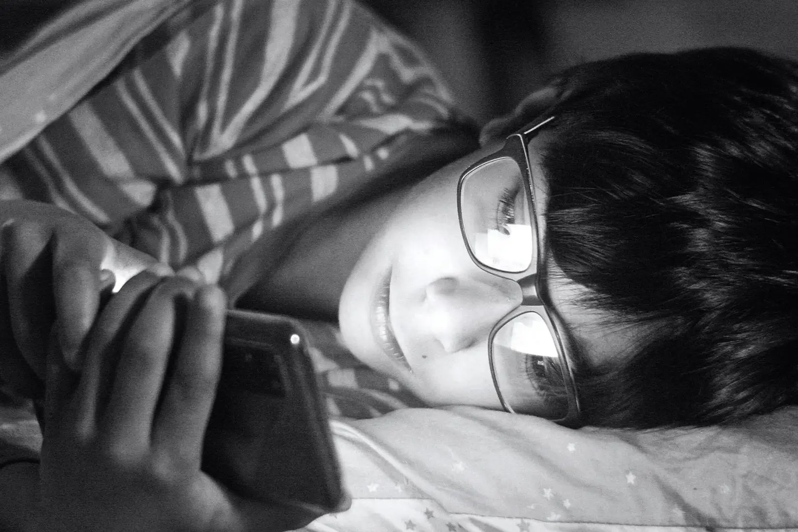 A boy wearing glasses lies in bed, holding a cell phone and looking at the screen with a focused expression.