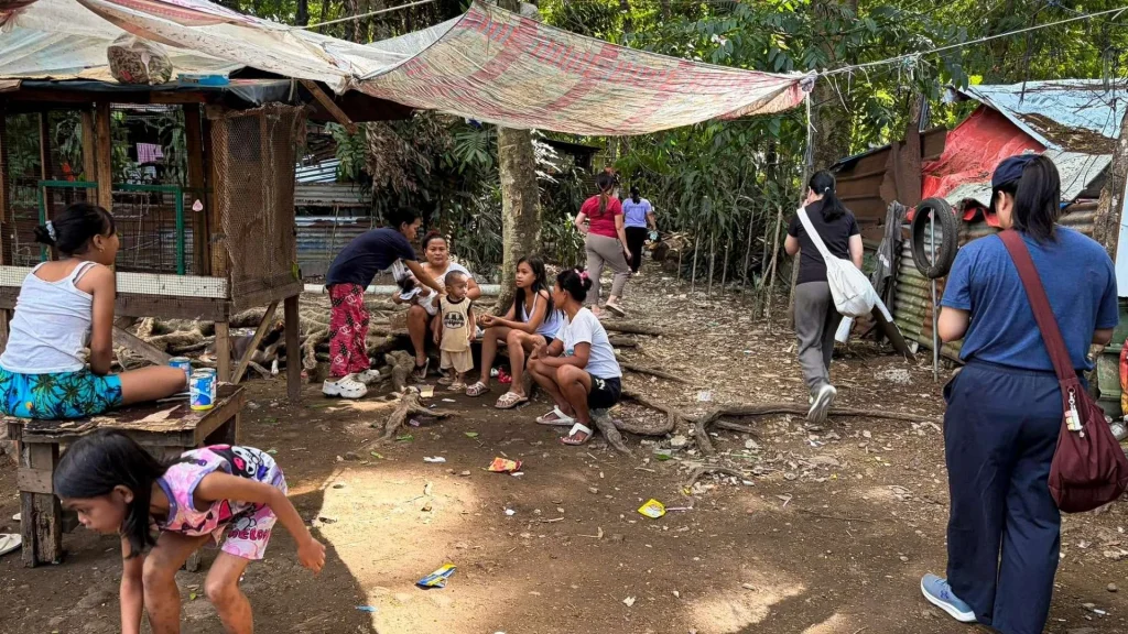 People sitting on the ground near a hut in a densely forested slum area in the Philippines, surrounded by trees and foliage.