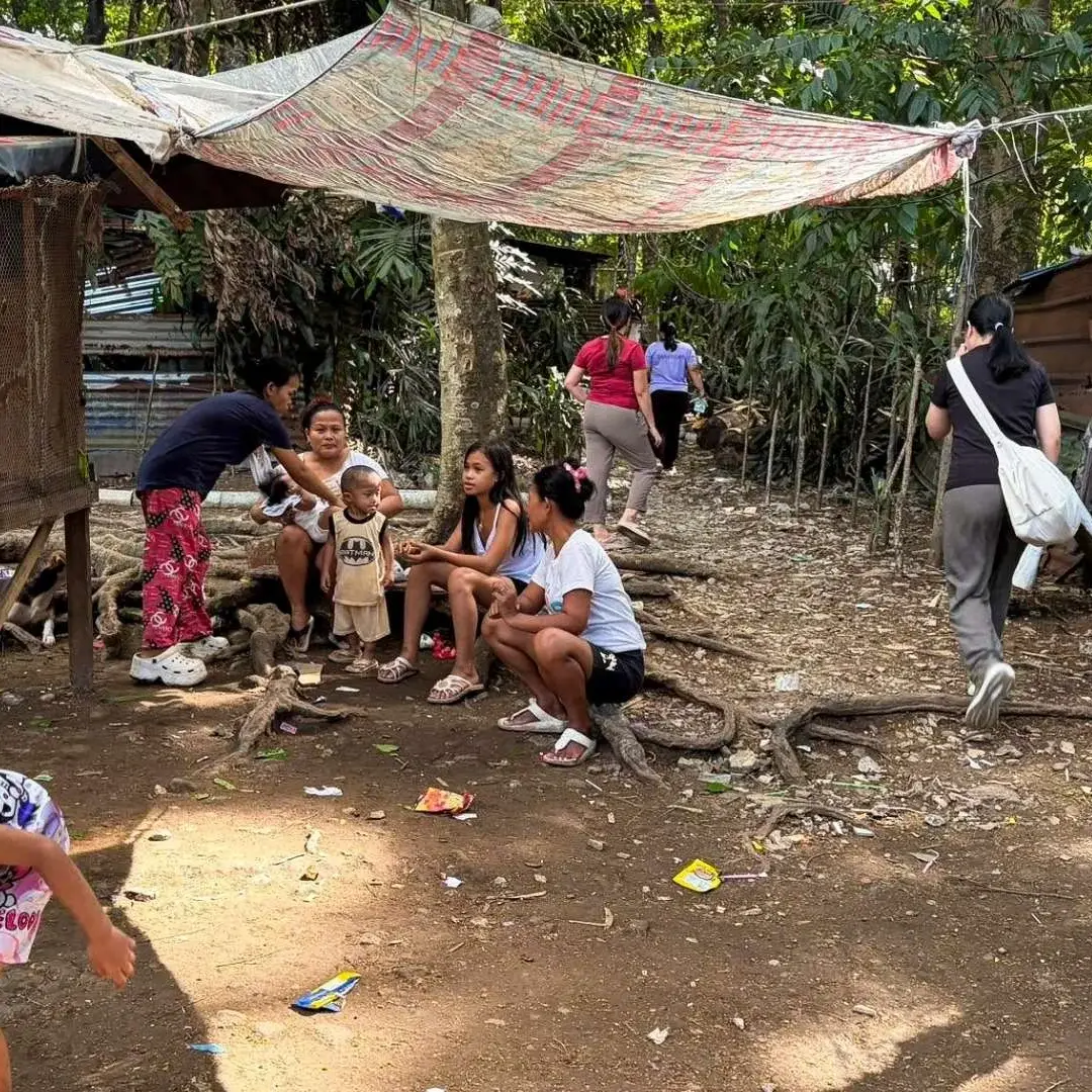 People sitting on the ground near a hut in a densely forested slum area in the Philippines, surrounded by trees and foliage.