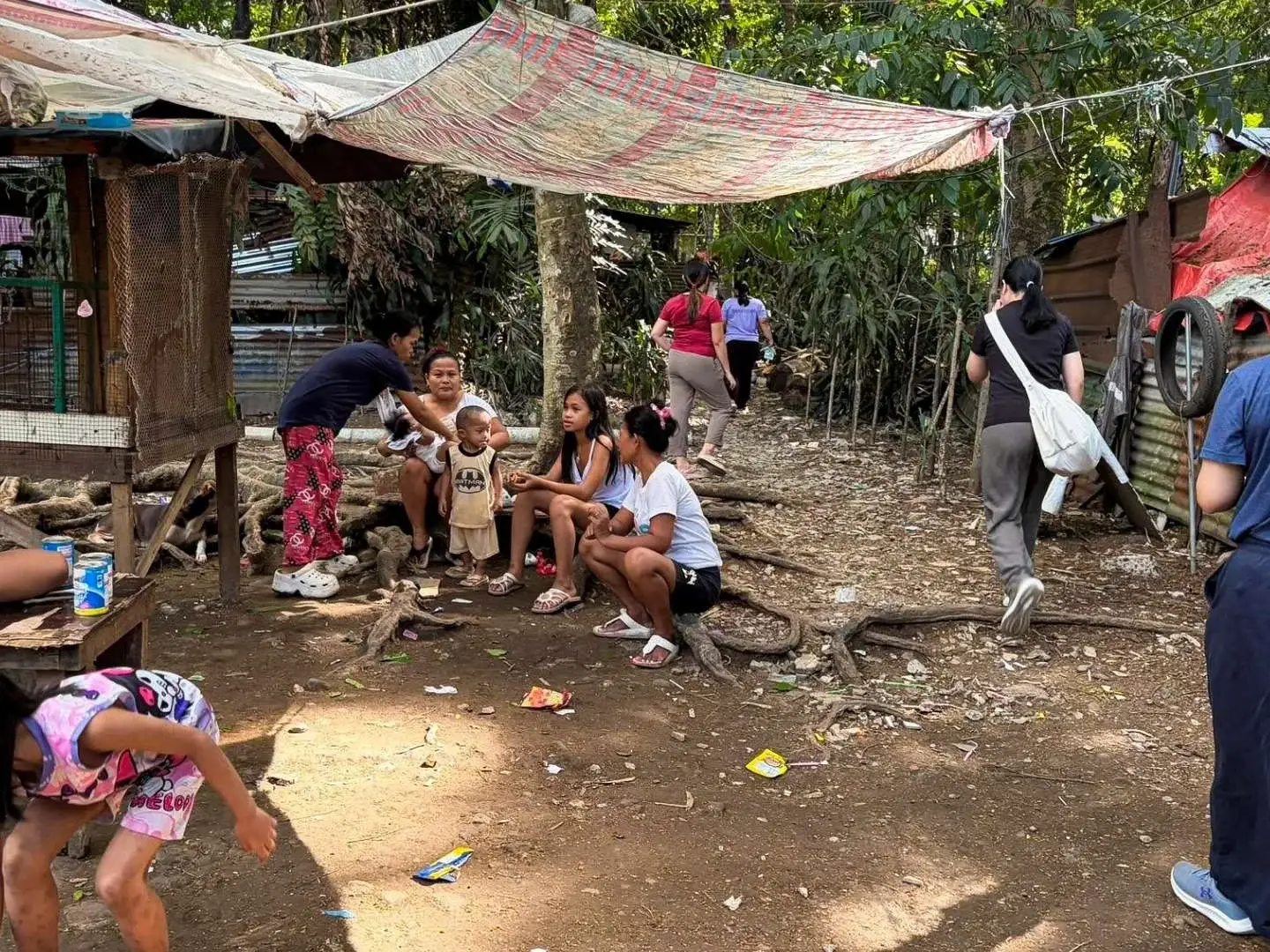 People sitting on the ground near a hut in a densely forested slum area in the Philippines, surrounded by trees and foliage.
