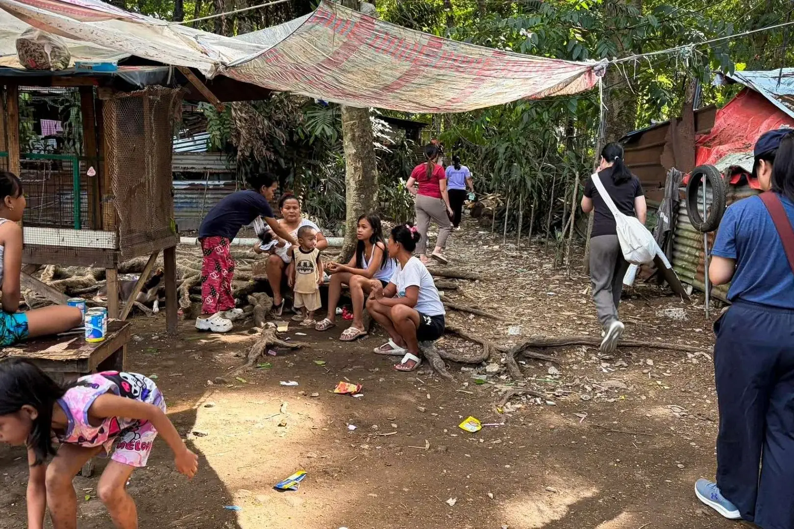 People sitting on the ground near a hut in a densely forested slum area in the Philippines, surrounded by trees and foliage.