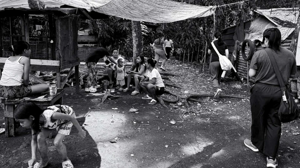 Black-and-white scene of children and adults in a crowded makeshift settlement, gathered beneath tarps and beside wooden shelters; the mood is stark and subdued.