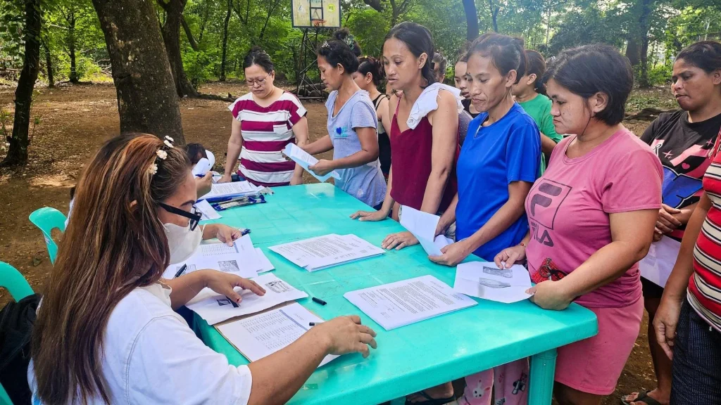 A woman at a table processes paperwork for a line of women standing outdoors in a park setting, with documents spread across the green table in the Philippines.