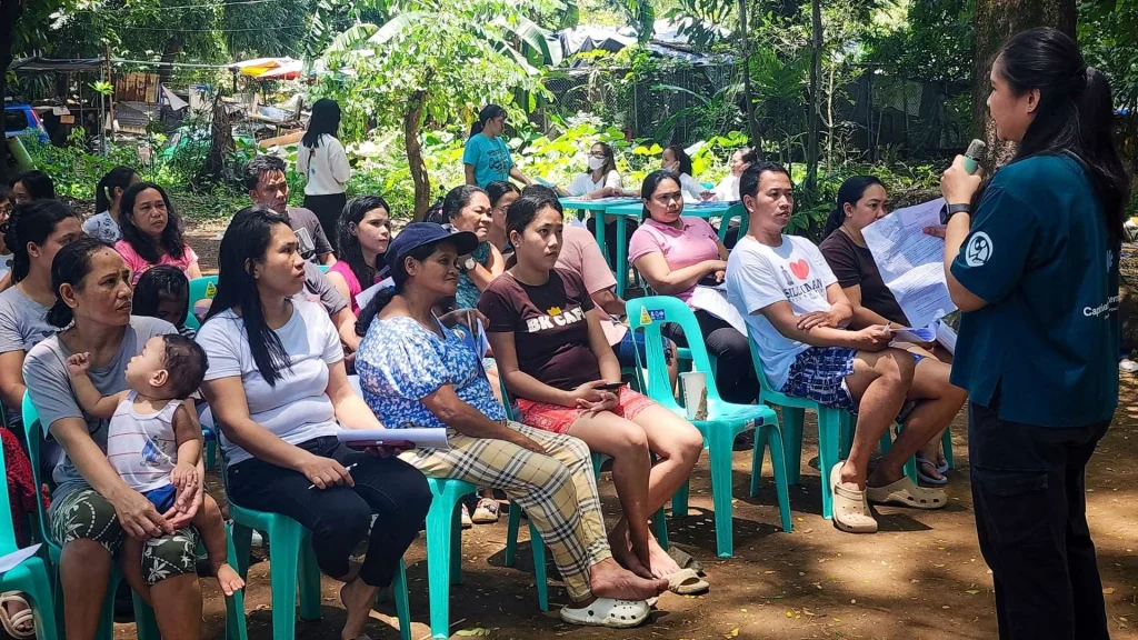 A woman speaks into a microphone to a group of people sitting on plastic chairs in an outdoor area in the Philippines, with trees and foliage in the background.
