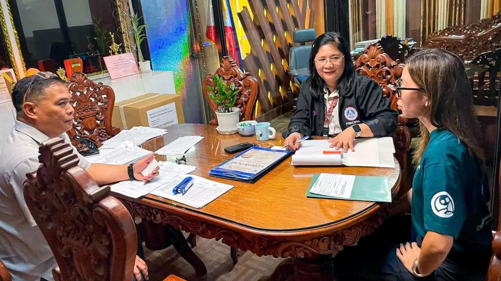 Three people sit around a wooden office table reviewing documents, with a woman in a black jacket and two others in casual shirts in a formal room in the Philippines.