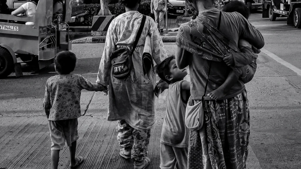Black-and-white street scene of adults walking with children, one carried on a hip and another holding a hand, amid traffic; candid, subdued mood.