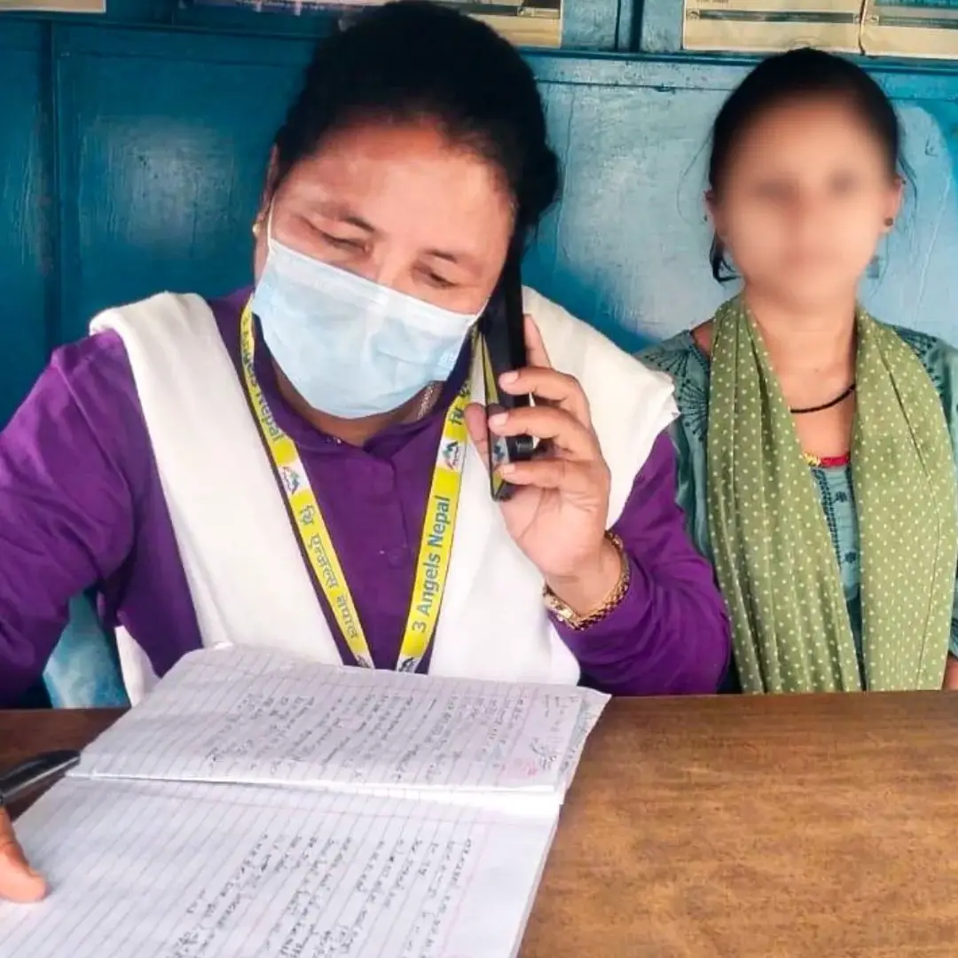 A woman wearing a mask talks on the phone while writing in a notebook. She is seated at a wooden table with a blurred person beside her, in a blue-walled room in Nepal.