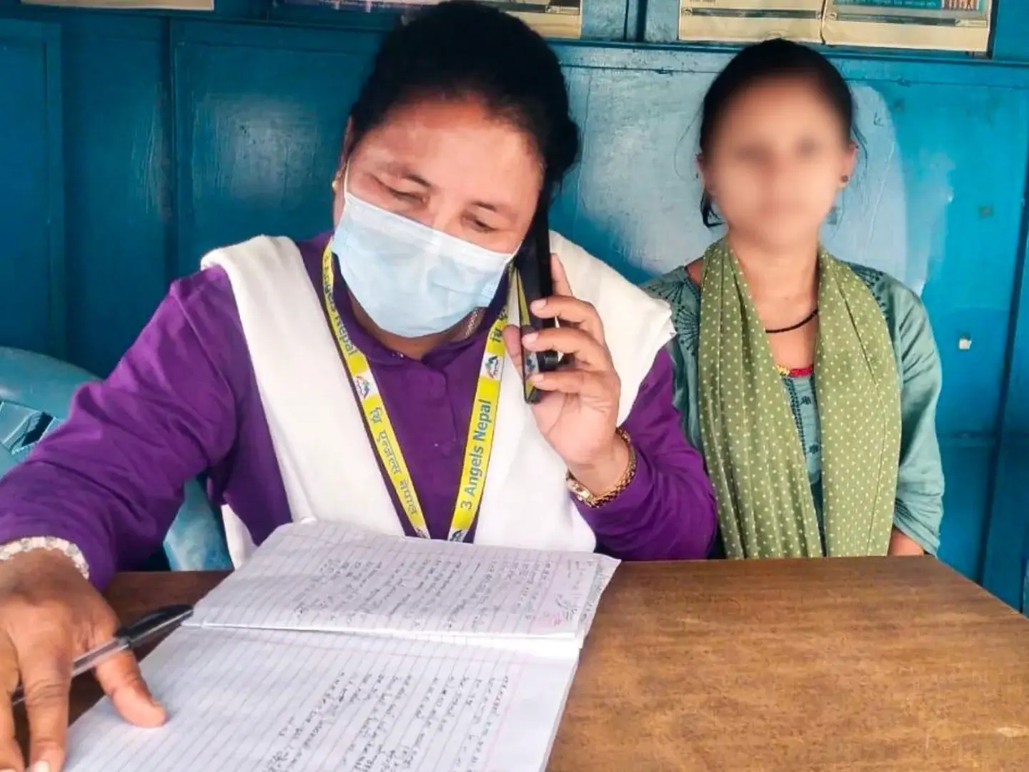 A woman wearing a mask talks on the phone while writing in a notebook. She is seated at a wooden table with a blurred person beside her, in a blue-walled room in Nepal.