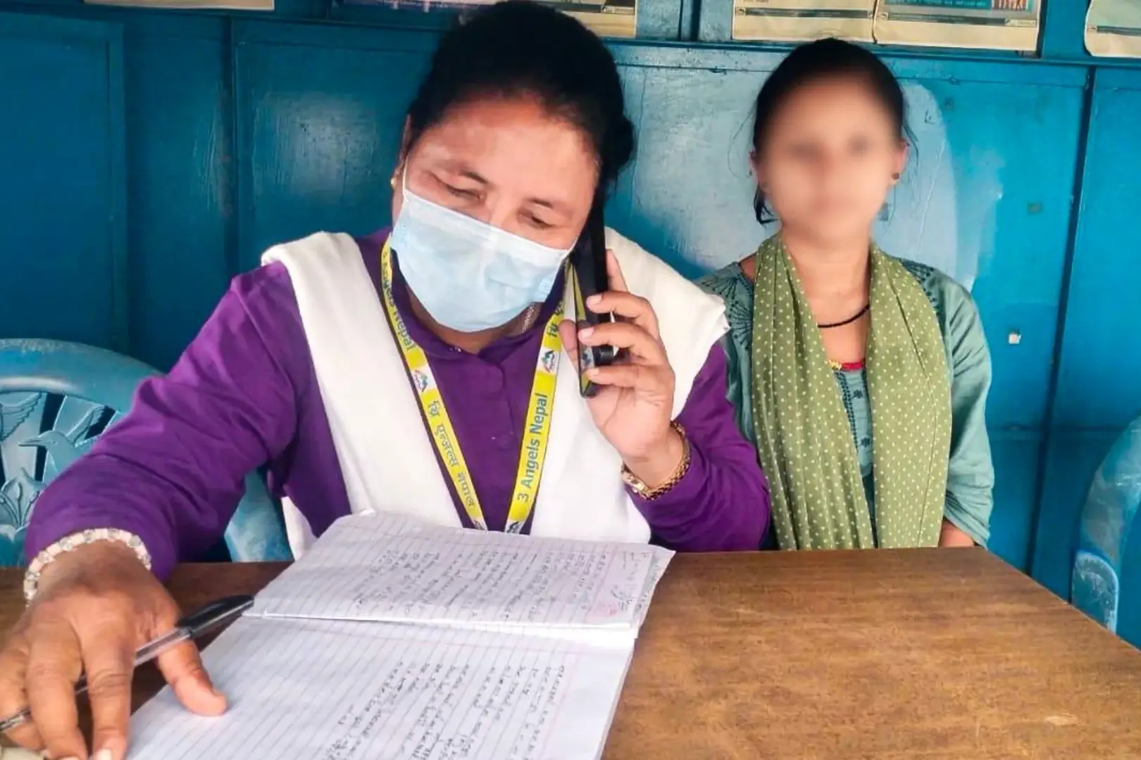 A woman wearing a mask talks on the phone while writing in a notebook. She is seated at a wooden table with a blurred person beside her, in a blue-walled room in Nepal.