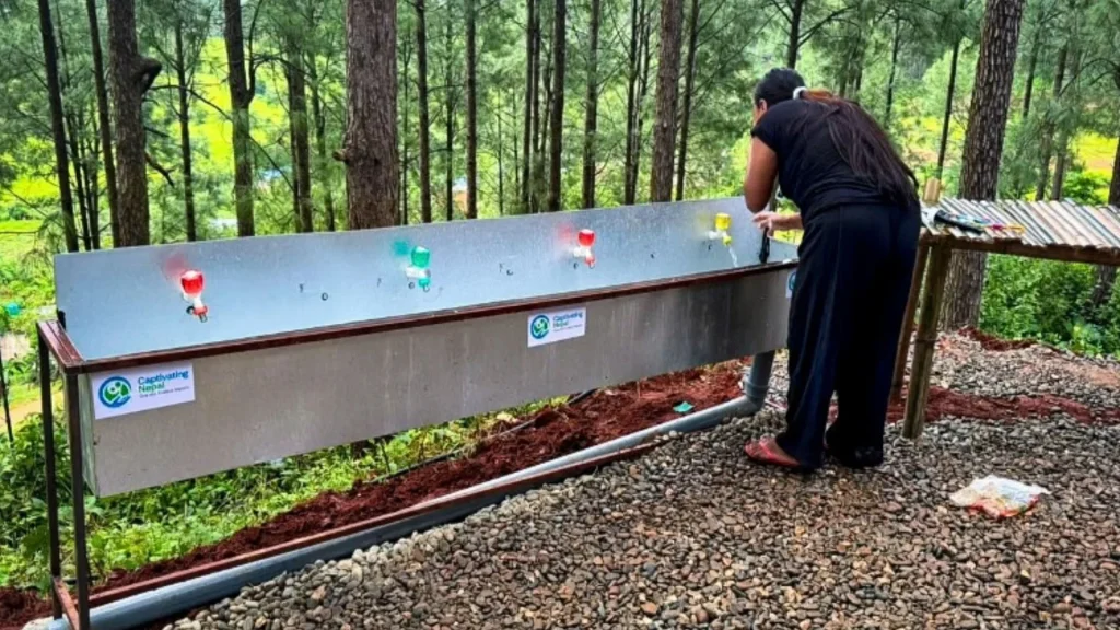 A woman in black stands by a long metal outdoor sink with colorful faucets in a forest, with Captivating Nepal labels on the side of the basin.