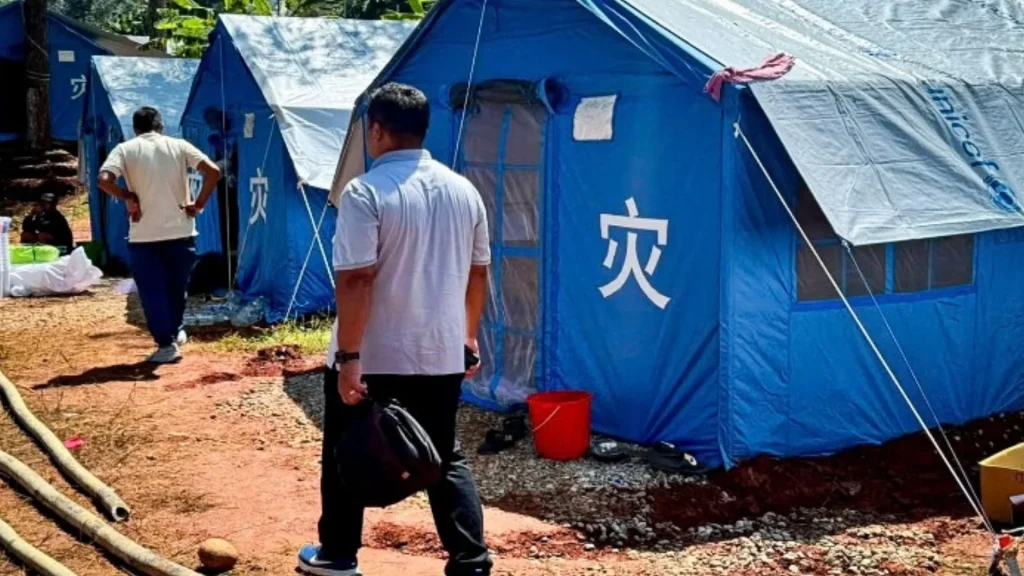 Two men walk past a row of blue emergency relief tents set up on a dirt ground, with a red bucket placed near the entrance of one tent in Nepal.