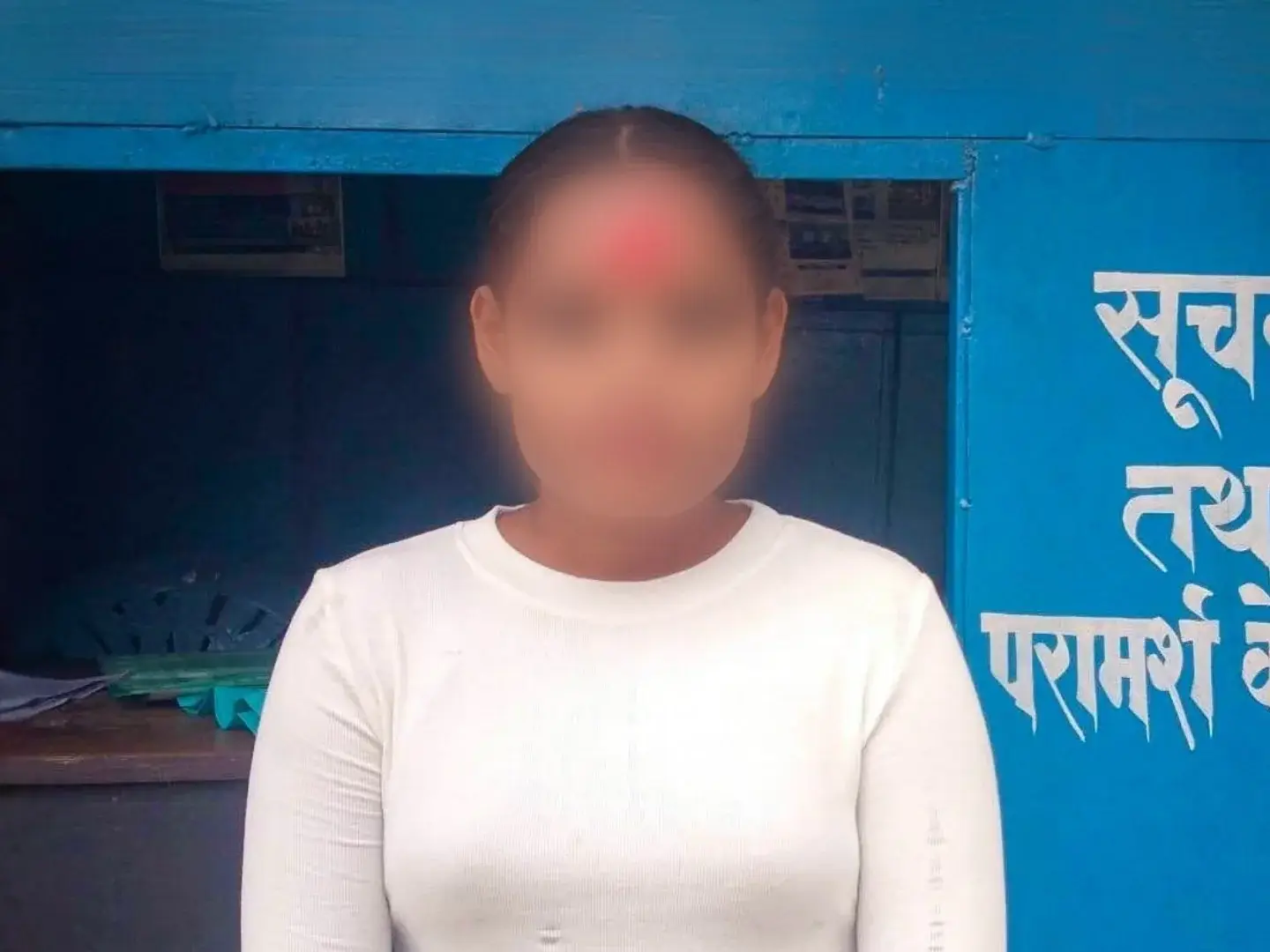 A blurred-faced person in a white long-sleeve shirt stands outside a bright blue Hindi-language information booth in Nepal, conveying a formal tone.