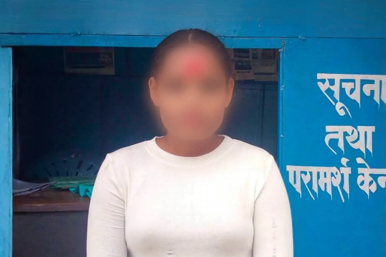 A blurred-faced person in a white long-sleeve shirt stands outside a bright blue Hindi-language information booth in Nepal, conveying a formal tone.