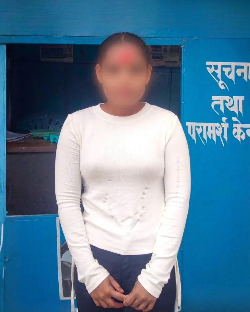 A blurred-faced person in a white long-sleeve shirt stands outside a bright blue Hindi-language information booth in Nepal, conveying a formal tone.