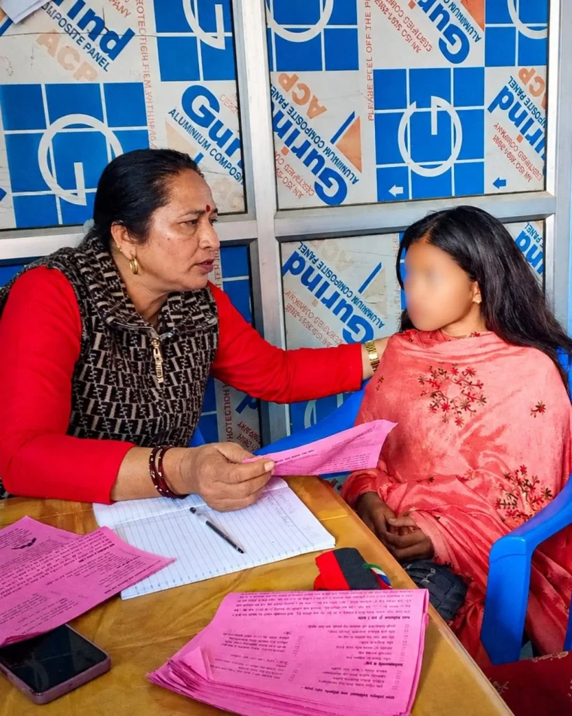 Woman in red offers a pink form to a child in pink at a paperwork-filled table, seated in blue chairs against a blue-and-white backdrop in Nepal. The girl's face is blurred for privacy.