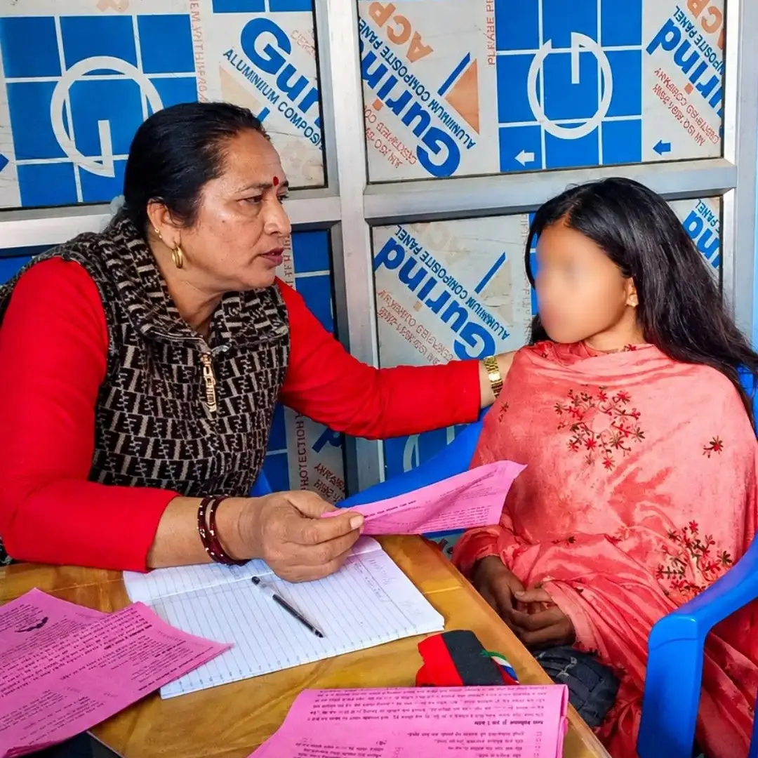Woman in red offers a pink form to a child in pink at a paperwork-filled table, seated in blue chairs against a blue-and-white backdrop in Nepal. The girl's face is blurred for privacy.