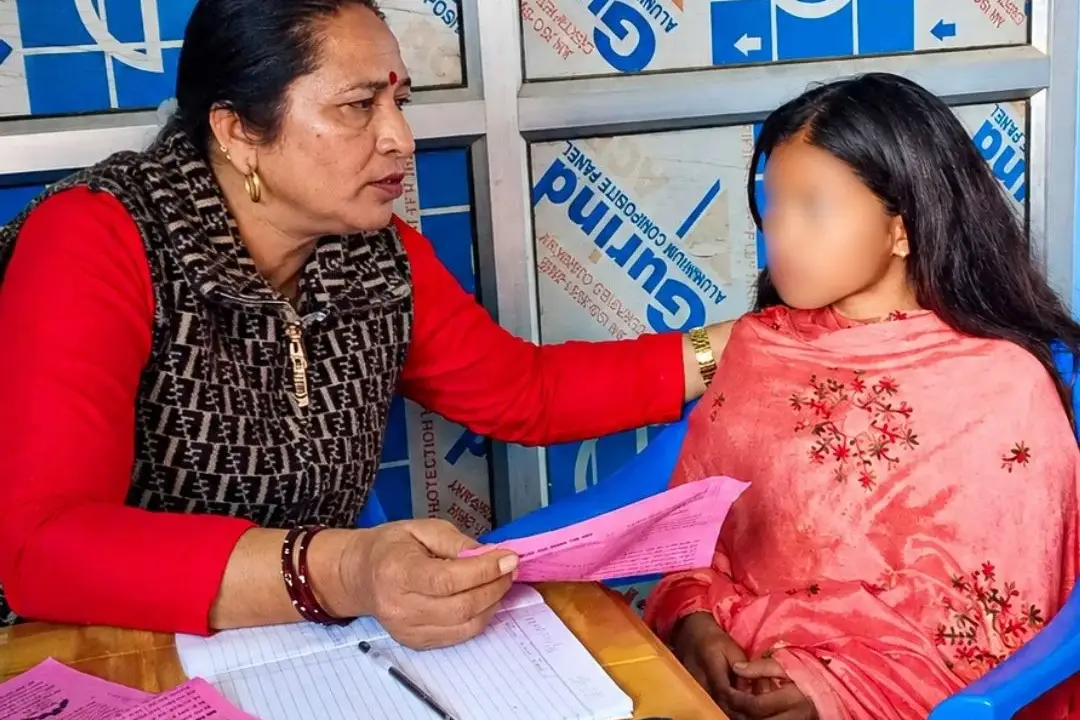 Woman in red offers a pink form to a child in pink at a paperwork-filled table, seated in blue chairs against a blue-and-white backdrop in Nepal. The girl's face is blurred for privacy.