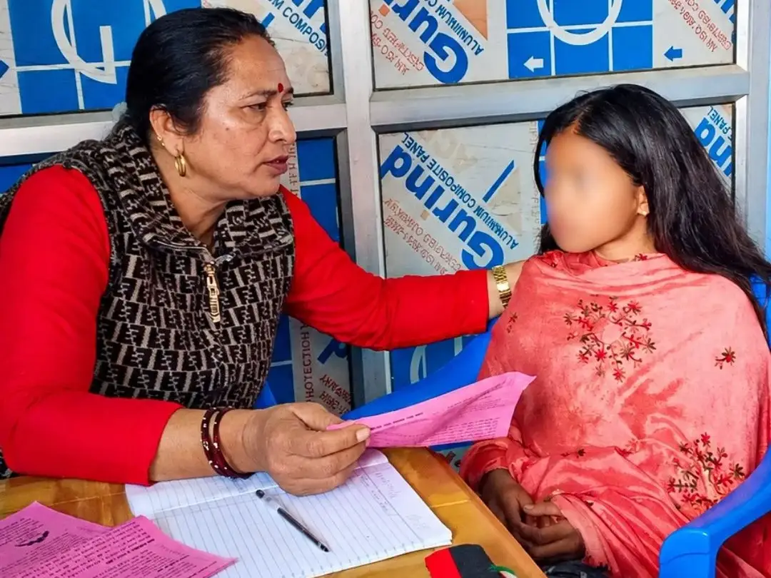 Woman in red offers a pink form to a child in pink at a paperwork-filled table, seated in blue chairs against a blue-and-white backdrop in Nepal. The girl's face is blurred for privacy.