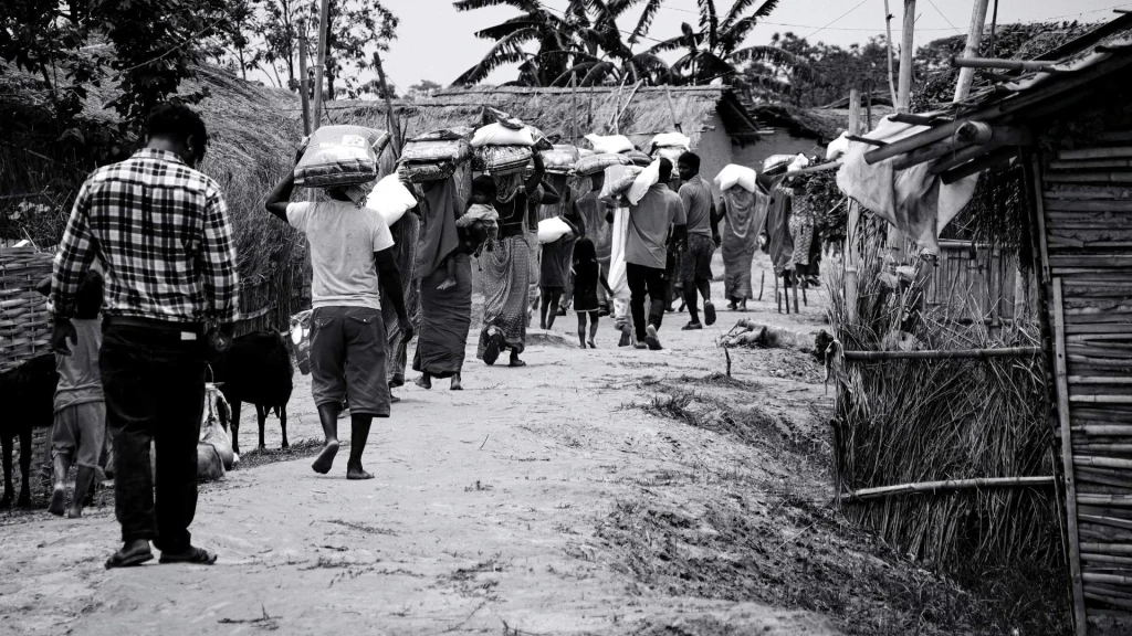 Black-and-white photo of villagers carrying bundles along a rural dirt road past thatched huts, conveying a somber, determined procession.